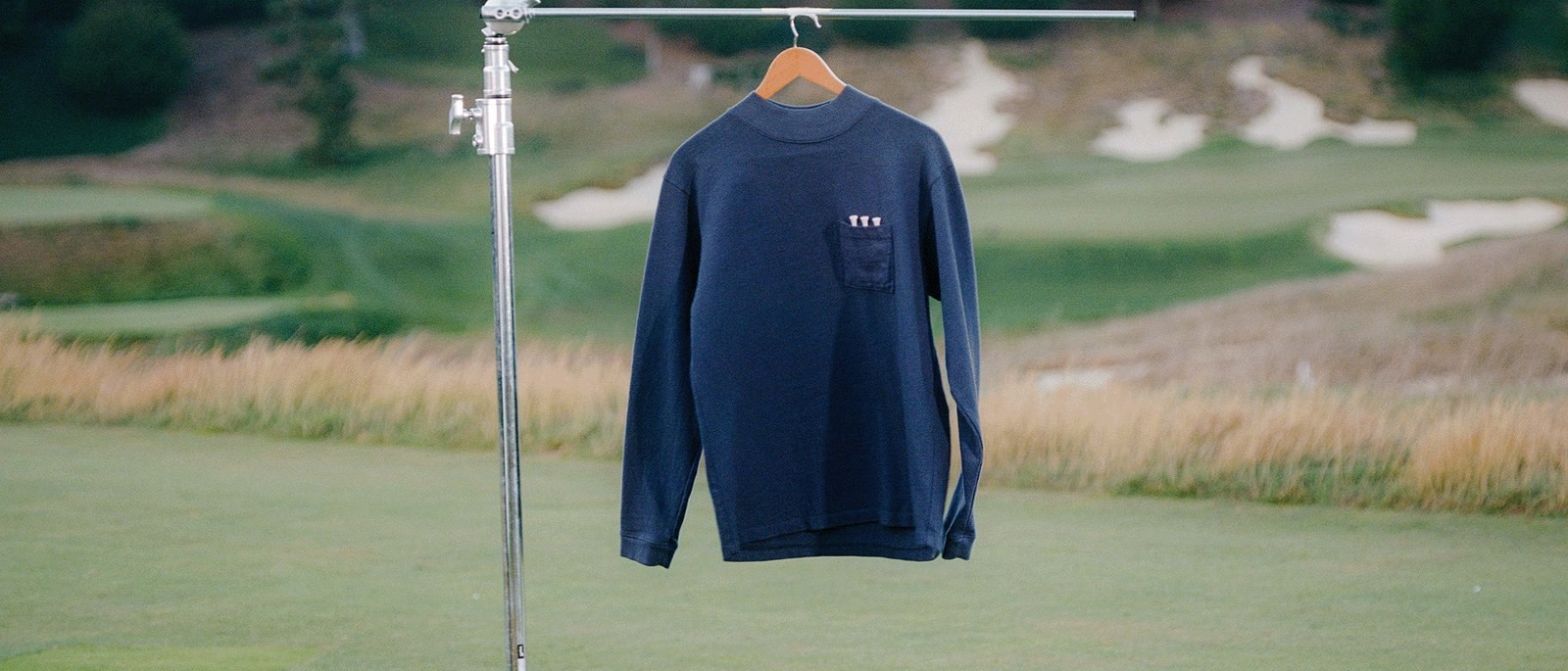 The Agronomy Workshop L/S Heavyweight Work Shirt in navy, hung on a garment rack in the middle of a golf course with three tees in the chest pocket