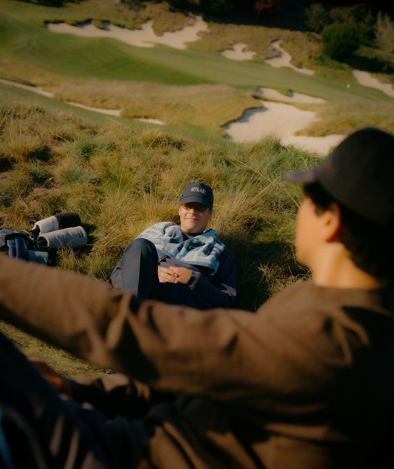 Two golfers sitting in the fescue above a bunkered links hole, one in the Work Shirt