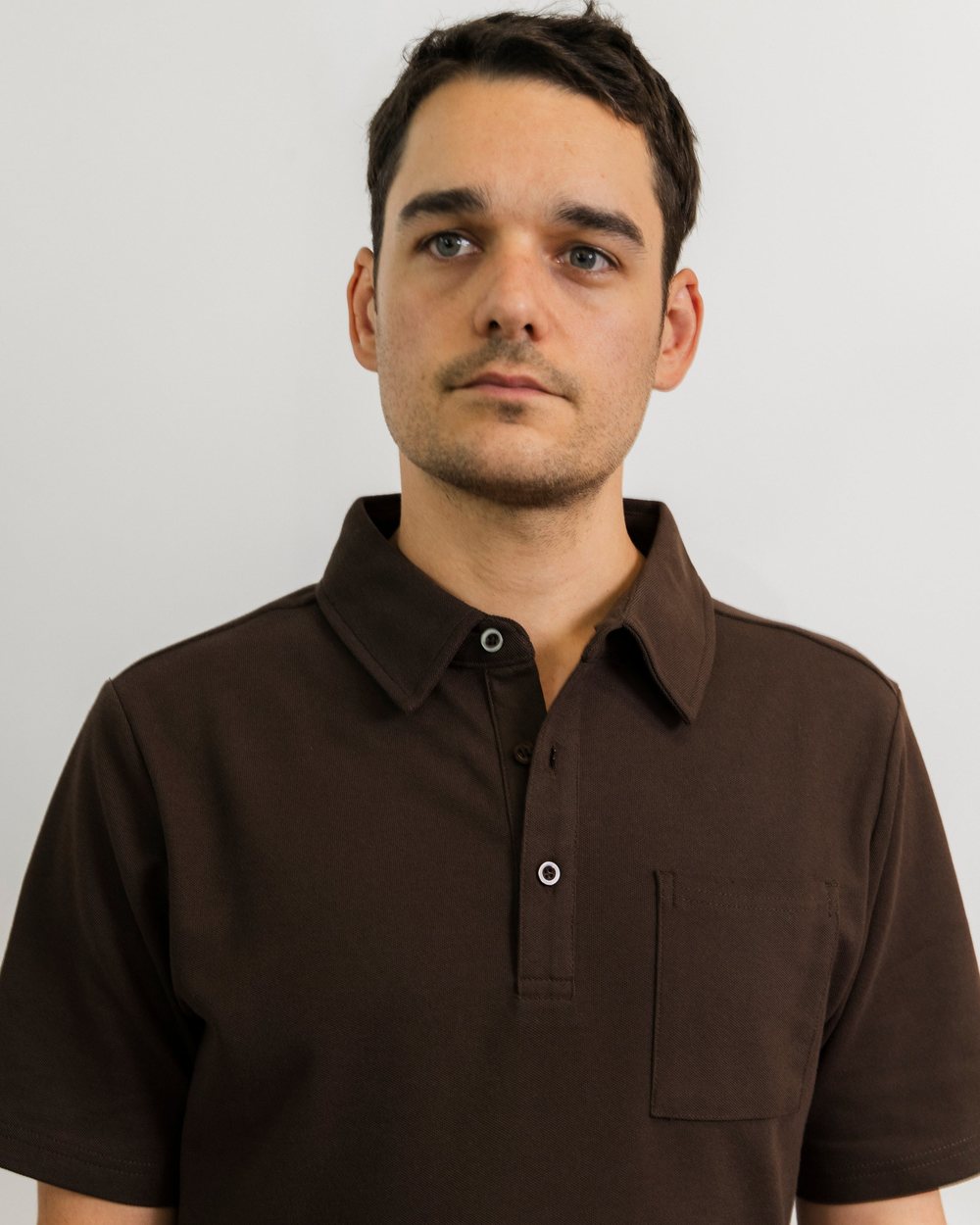 A man in a dark brown short sleeve pocket polo against a white wall