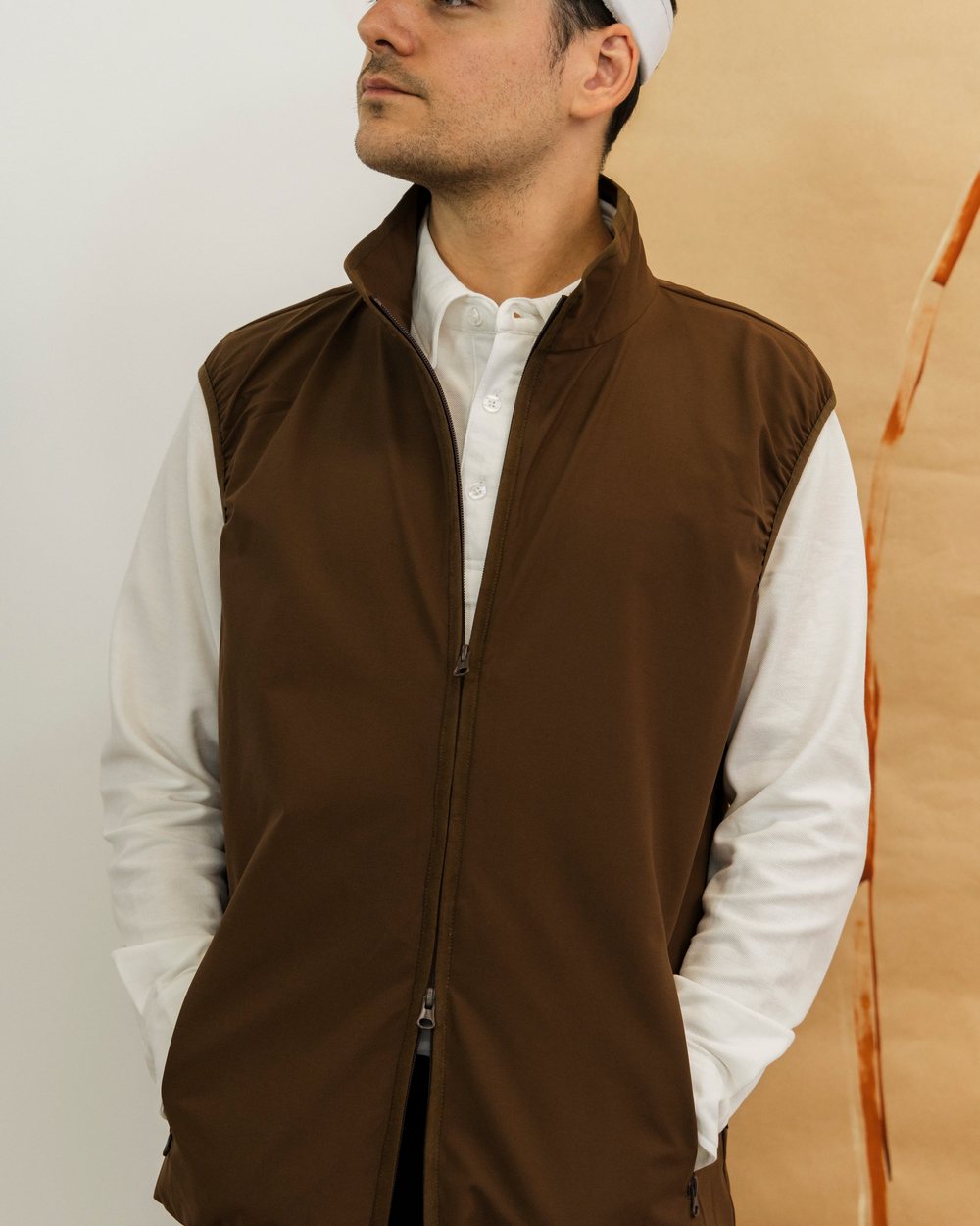 A man in a brown zip vest over a cream long sleeve polo and a white visor