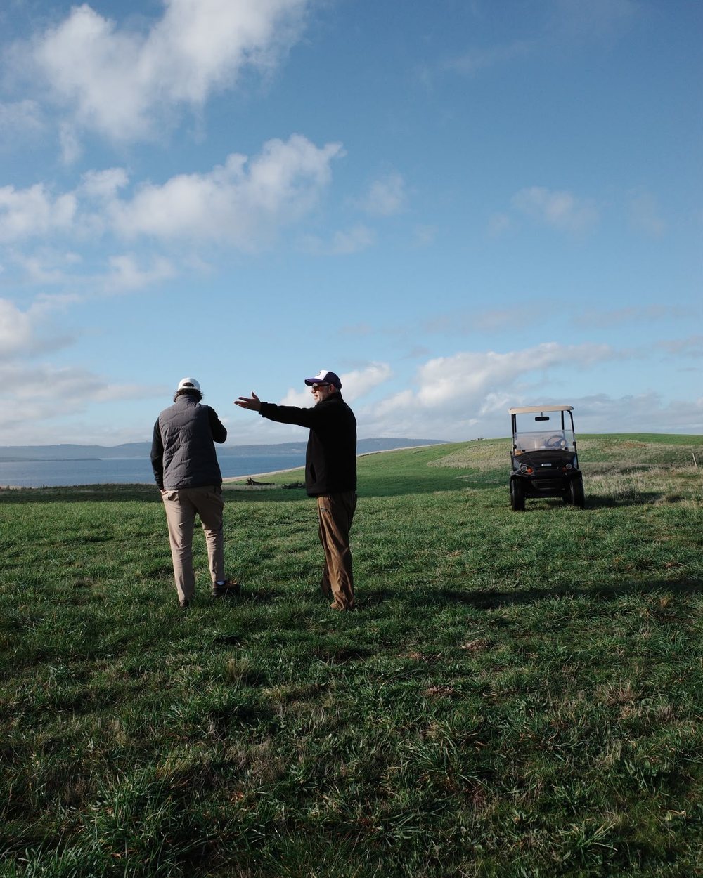 Two golfers on a wide green fairway pointing toward a cart under a big sky