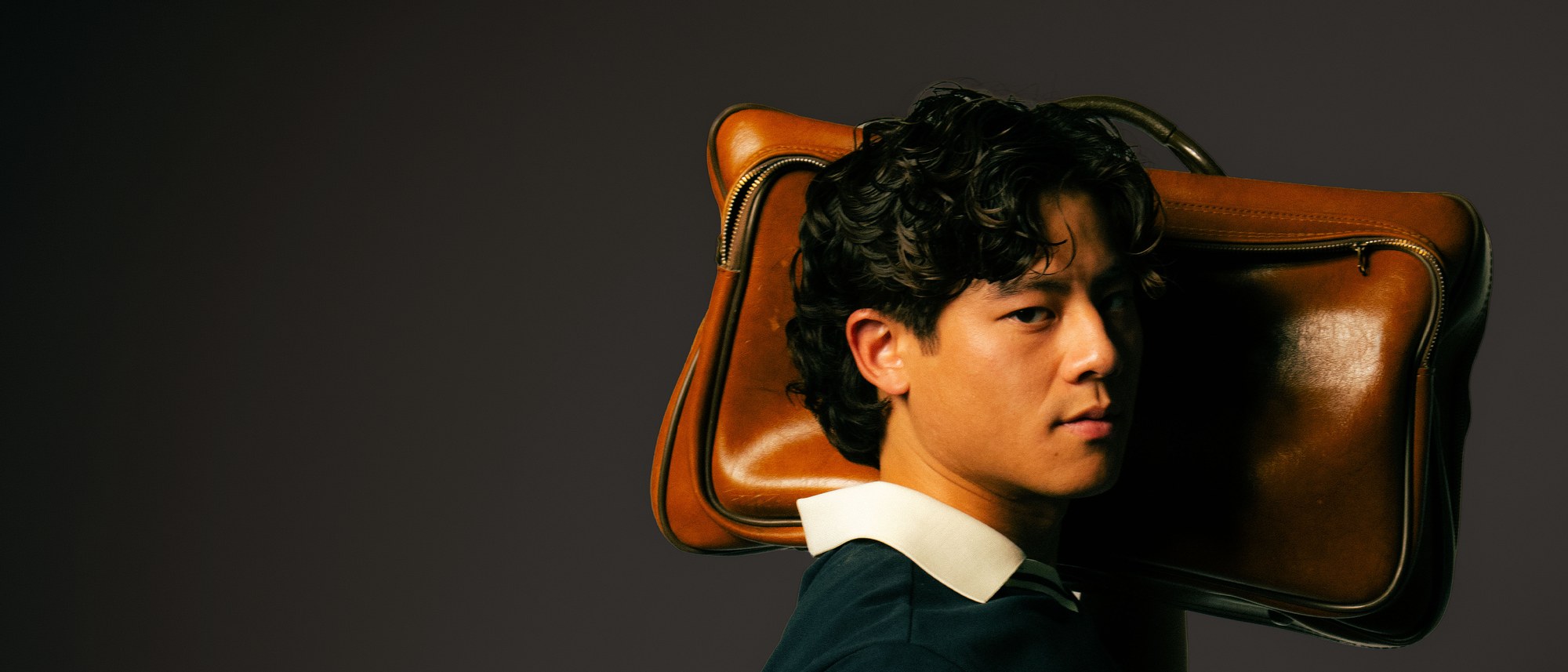Kingfisher Golf campaign photo: a man in a navy polo with a white collar carrying a vintage brown leather bag on his shoulder against a dark backdrop