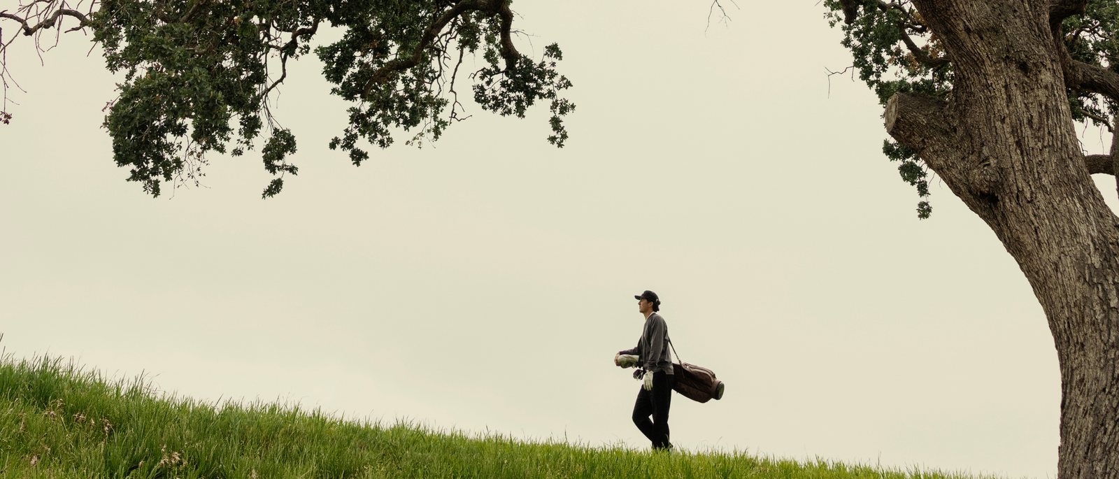 A golfer carrying his bag under a big oak on a hillside course, from Ashworth