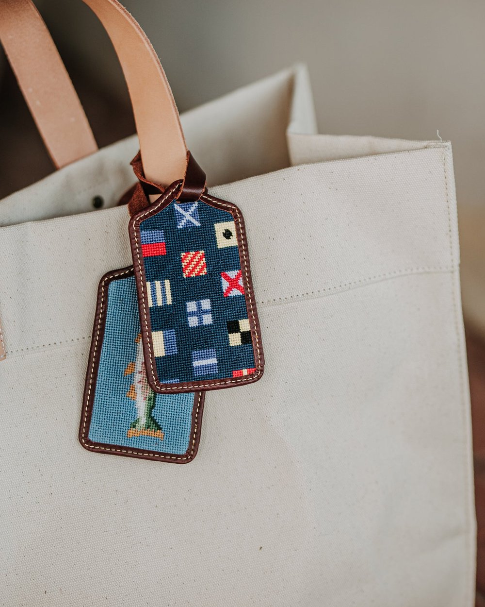 Two needlepoint Asher Riley luggage tags on a canvas tote