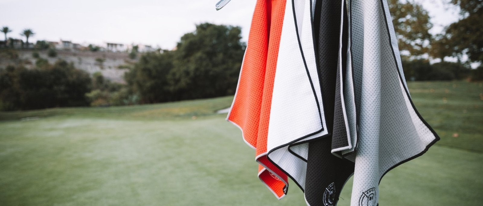 Four Ghost Golf towels in orange, white, black and grey hanging from a golf bag on the course