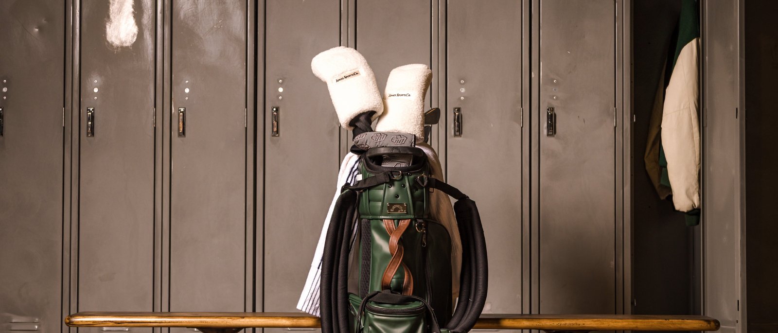 A green Jones golf bag with cream sherpa headcovers on a bench in front of steel lockers