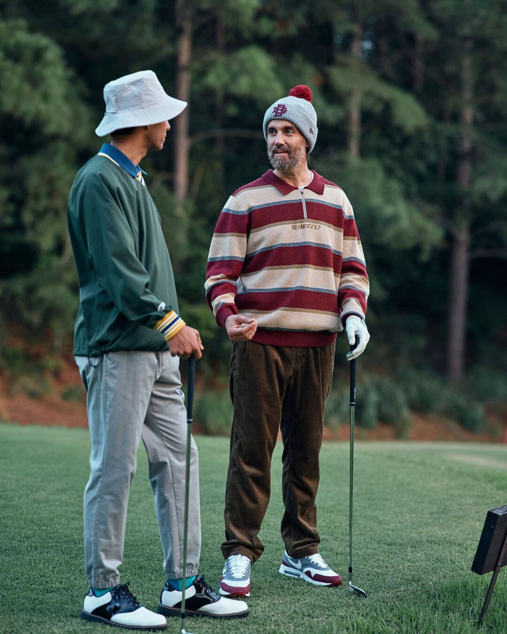 Two golfers talking on a tee, one in a green cardigan and bucket hat, one in a striped rugby knit and beanie