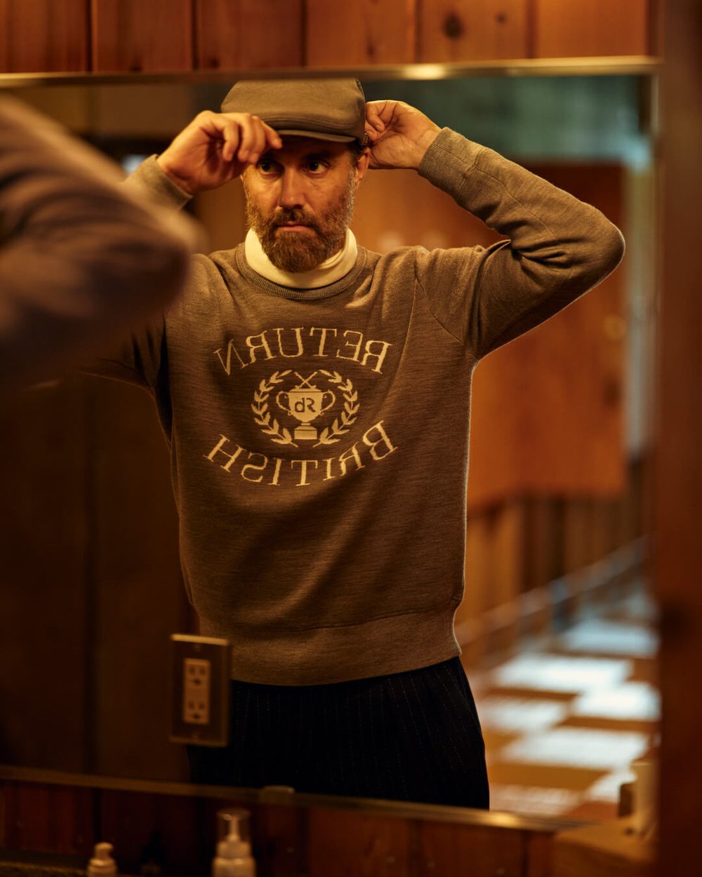 A bearded man in a Return British crest sweater adjusting a flat cap in a wood-paneled room