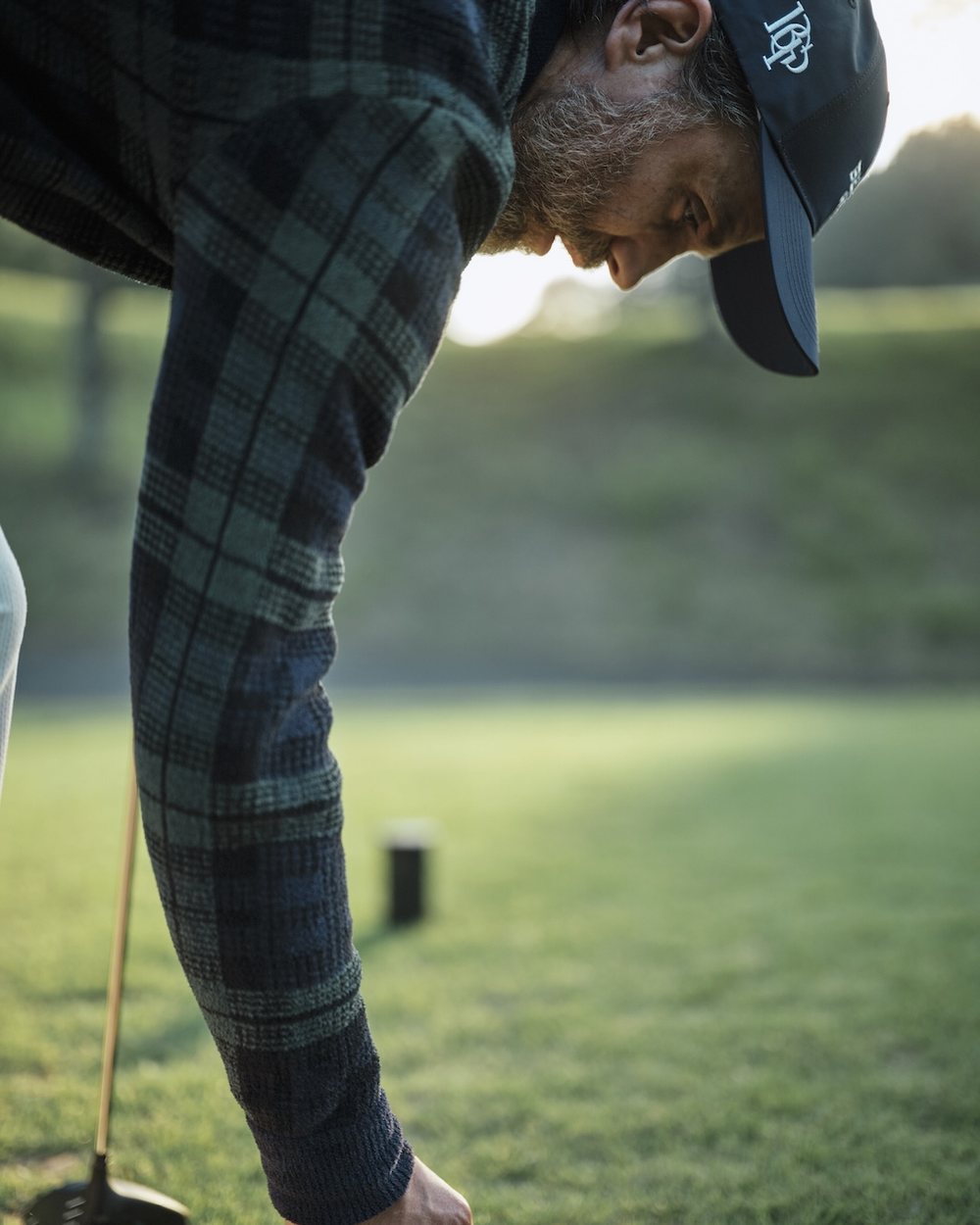A man in a tartan shirt and navy cap teeing up a ball