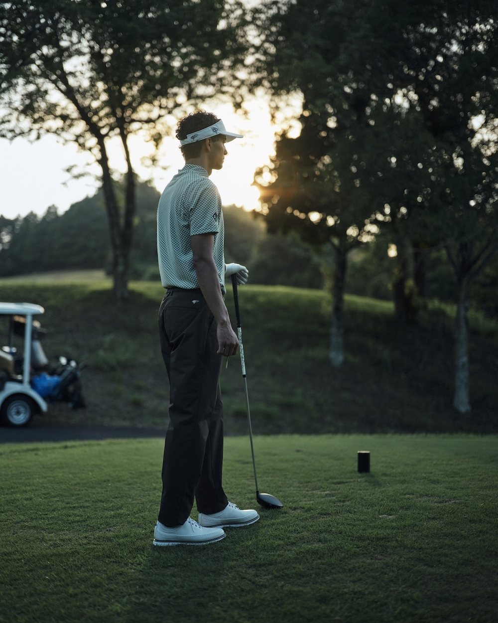 A golfer on the tee at sunset with a cart behind him