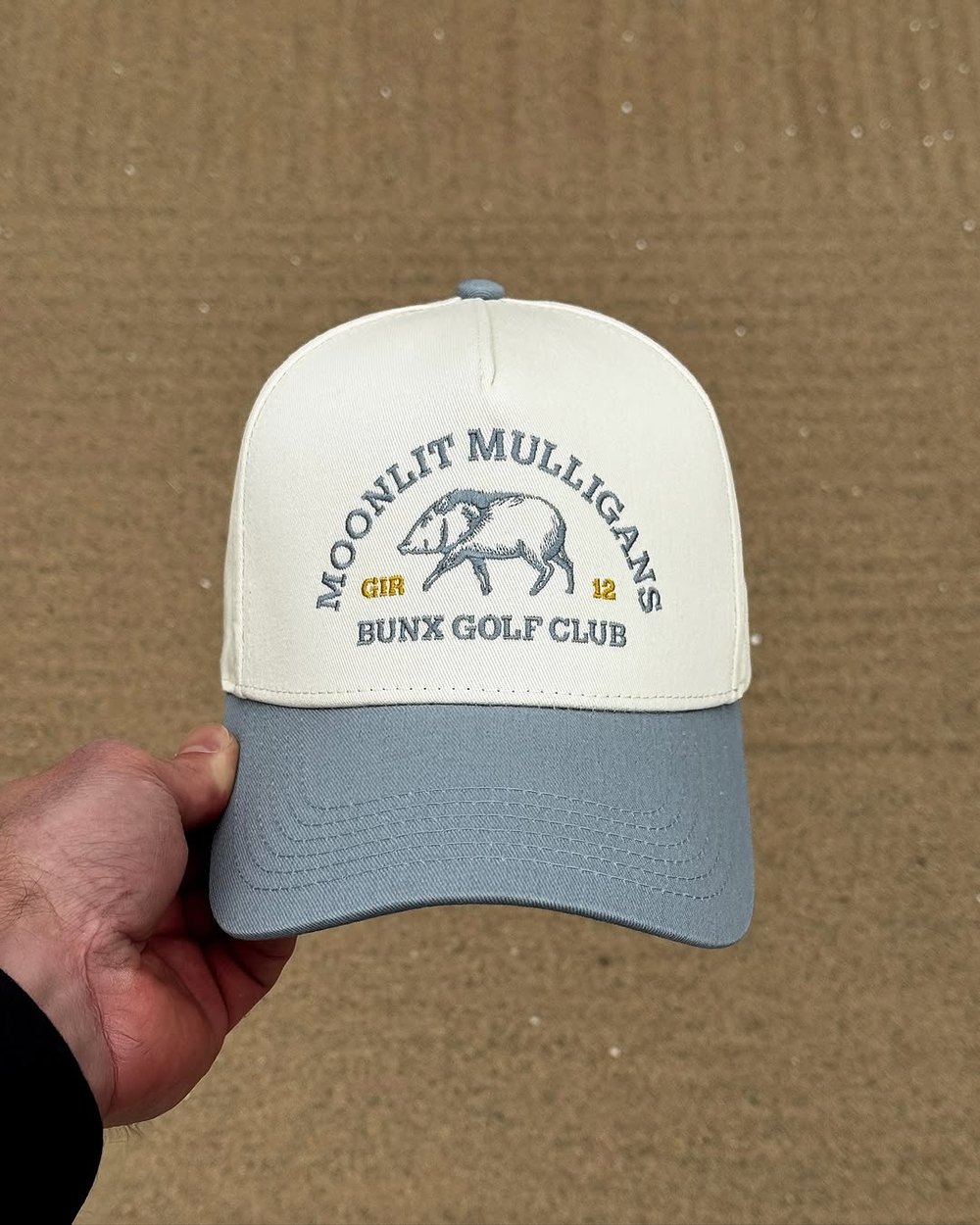 A hand holding the Moonlit Mulligans hat against a sand-colored wall