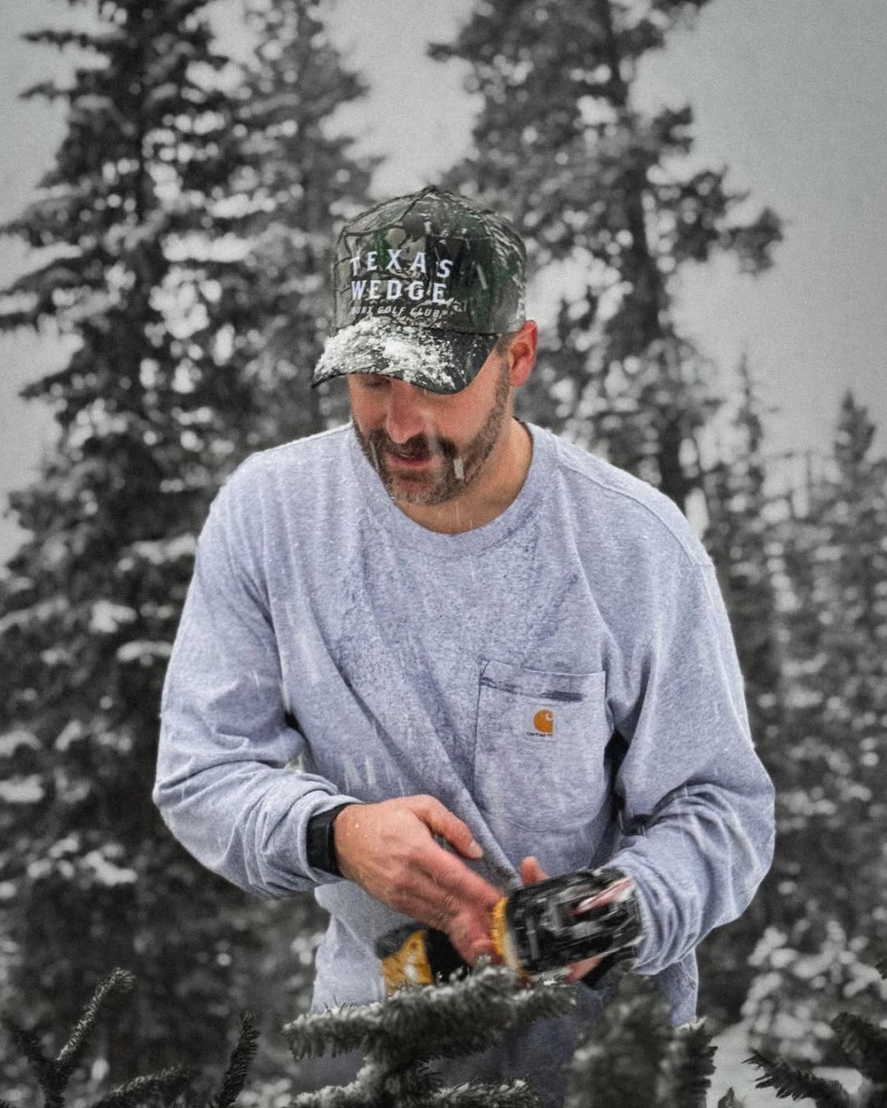 A man in a camo BUNX hat in a snowy pine forest
