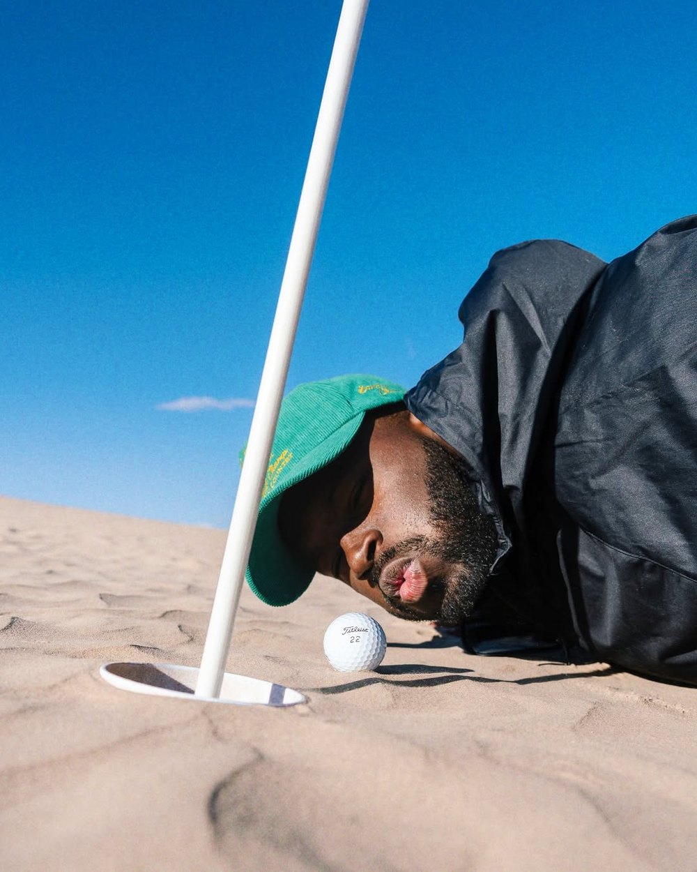 A golfer lying face-down in a bunker next to the flagstick and ball