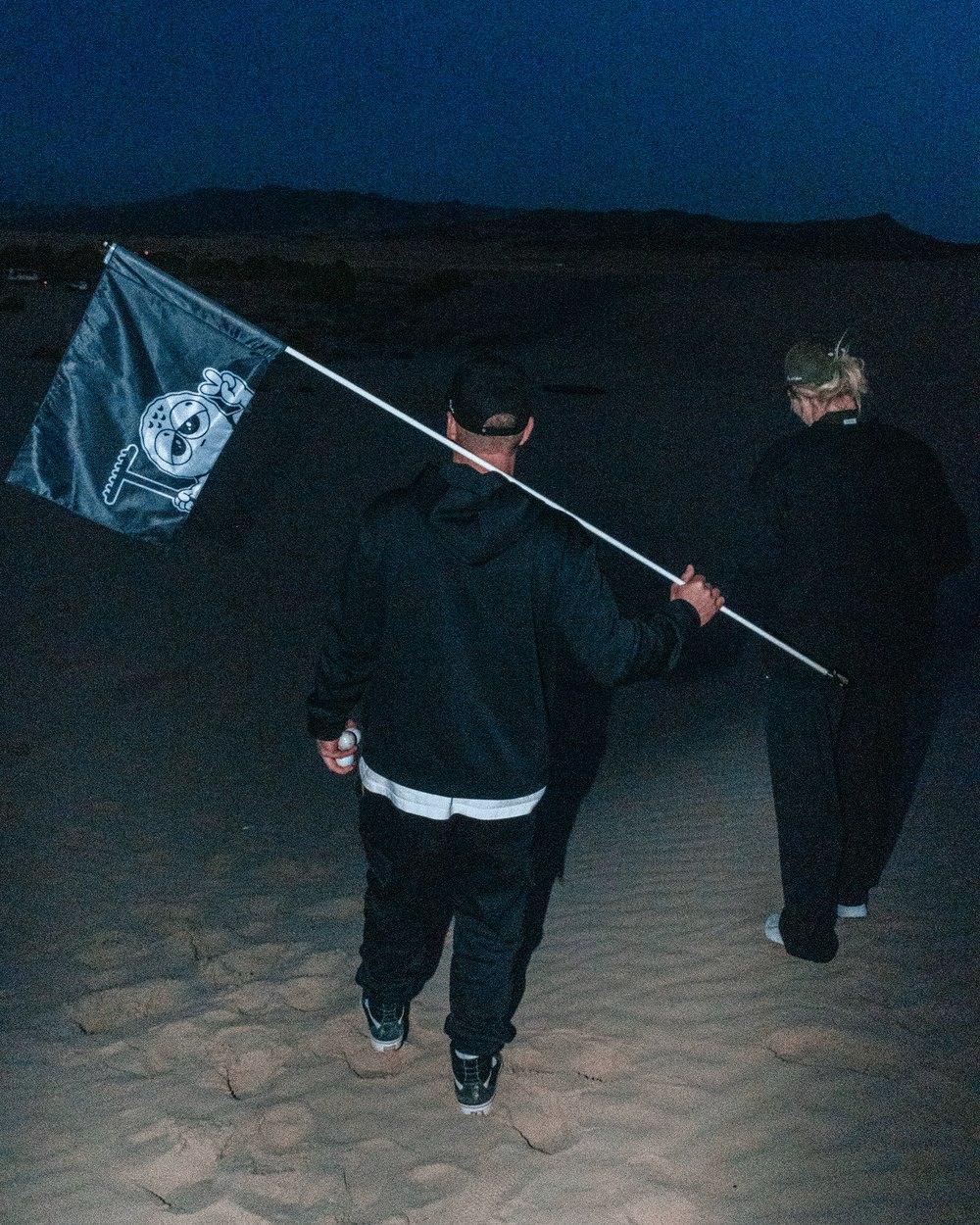 Two people walking across dunes at night carrying a BUNX flag