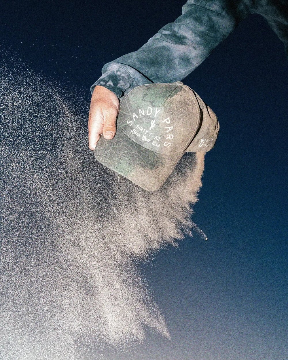 A hand pouring sand out of a camo Sandy Pars hat