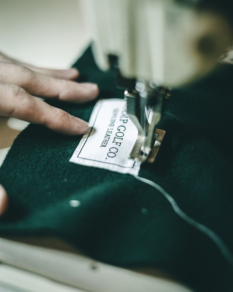 Hands sewing a Clamp Golf Co. label onto a green headcover