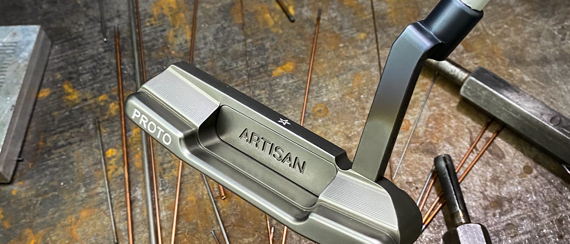 An Artisan Golf PROTO putter head resting on a steel workbench with copper rods and tools in the Fort Worth shop