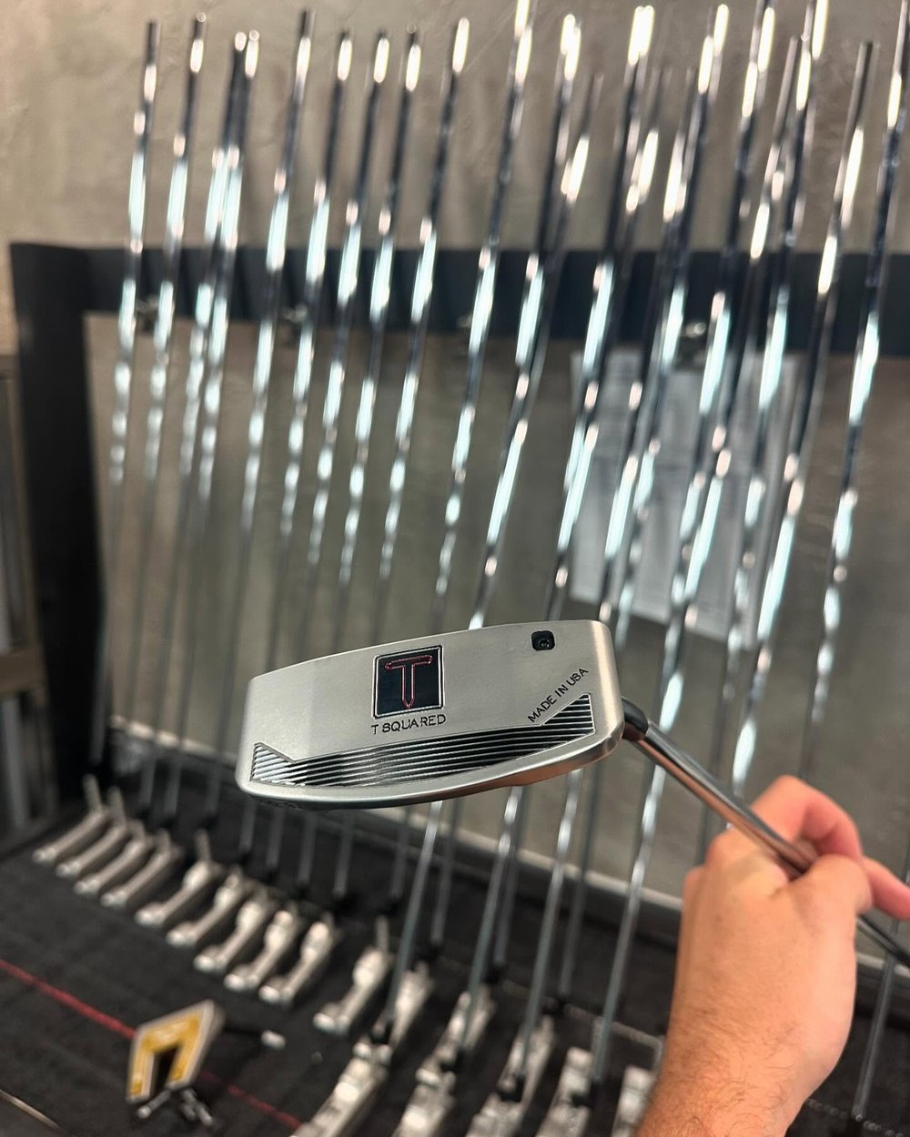 A T Squared putter held up in front of a rack of finished heads