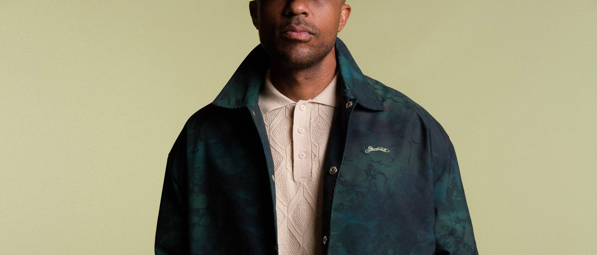 A model in a Students Golf dark green jacket over a cream cable-knit polo, against a pale sage background