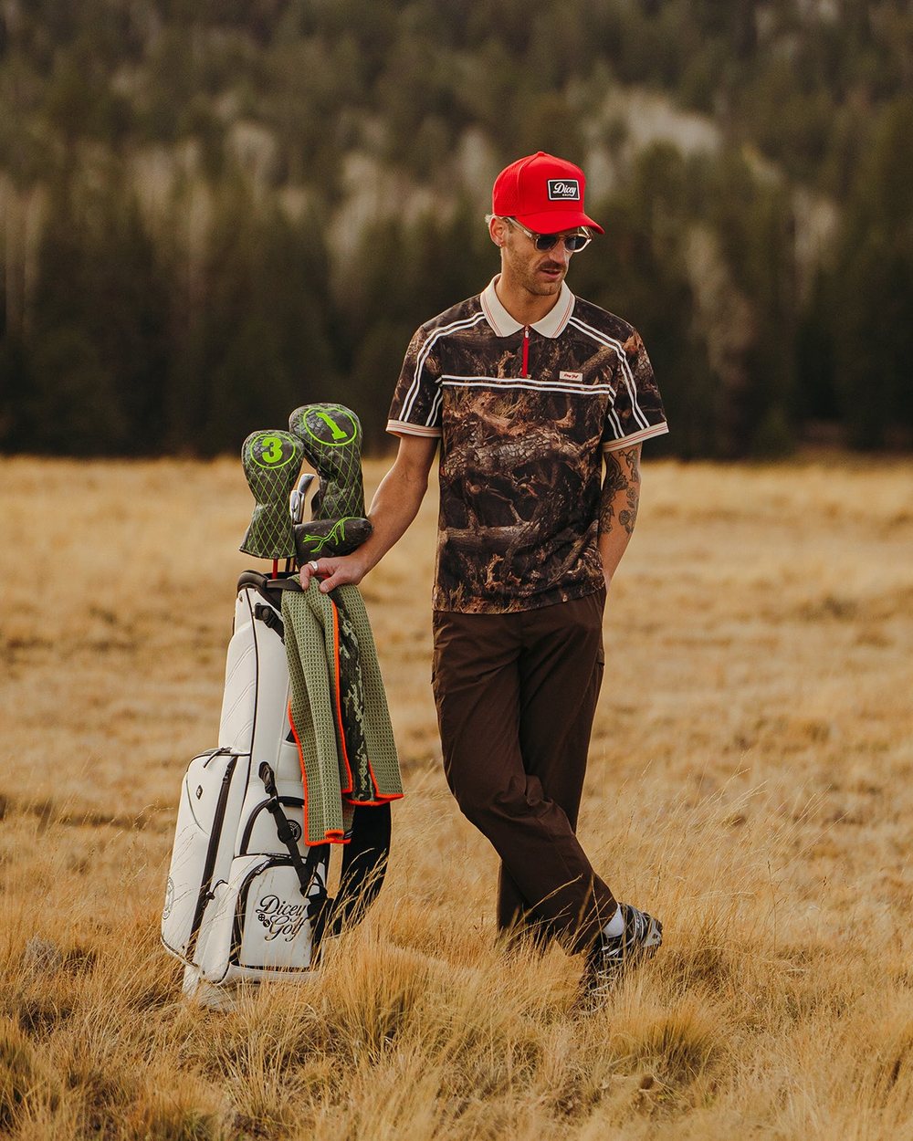 A golfer in a camo polo and red hat walking with his bag through dry grass