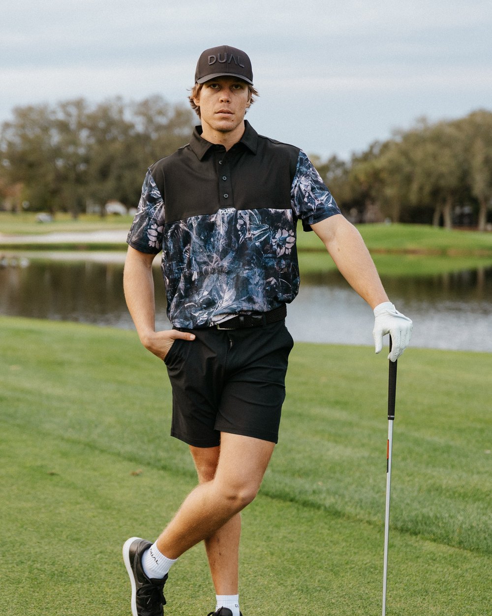 A golfer in the Midnight Bloom Signature polo and black shorts leaning on an iron beside a pond