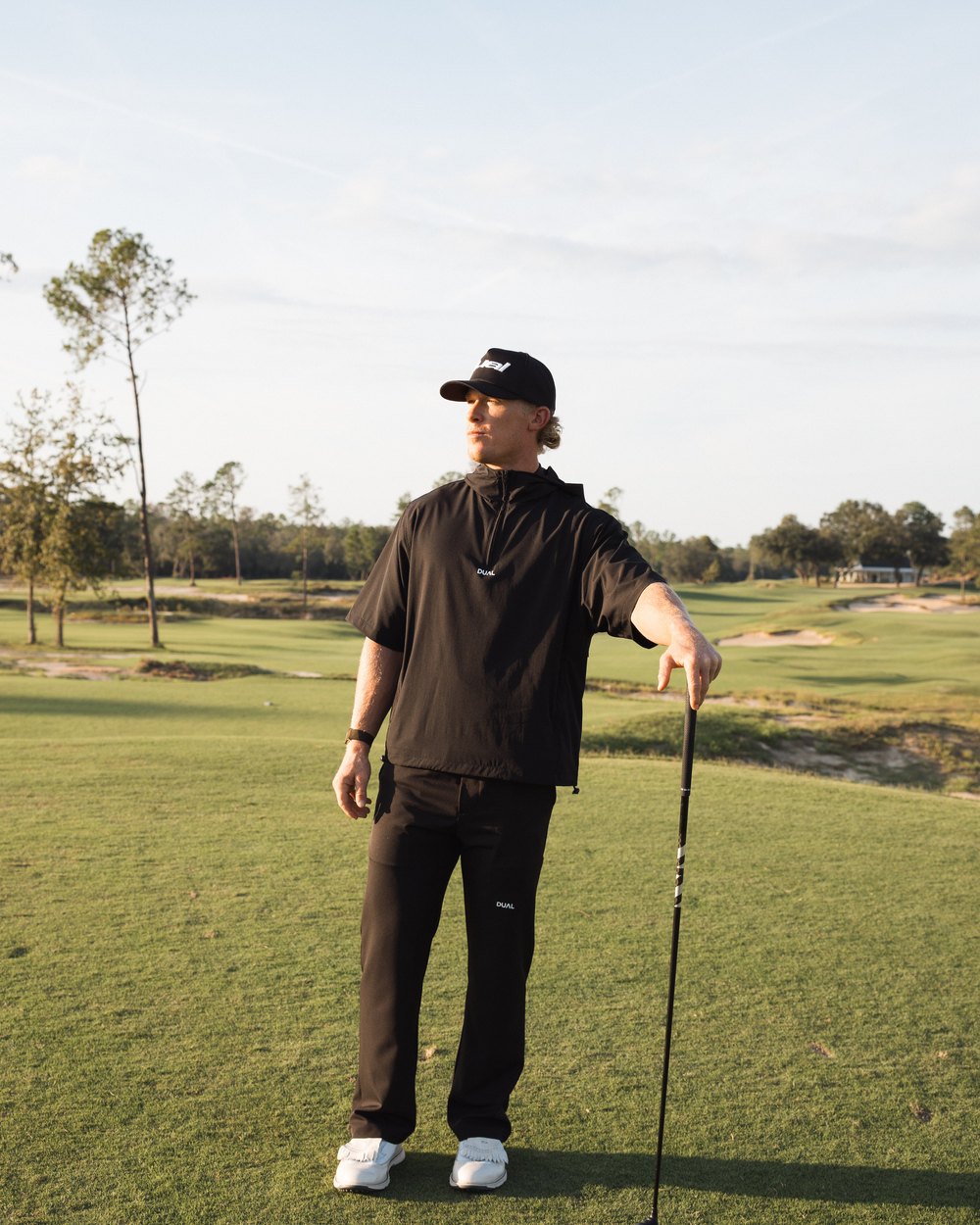 A golfer in the black Pursuit hooded windbreaker and black pants resting on a driver on the fairway