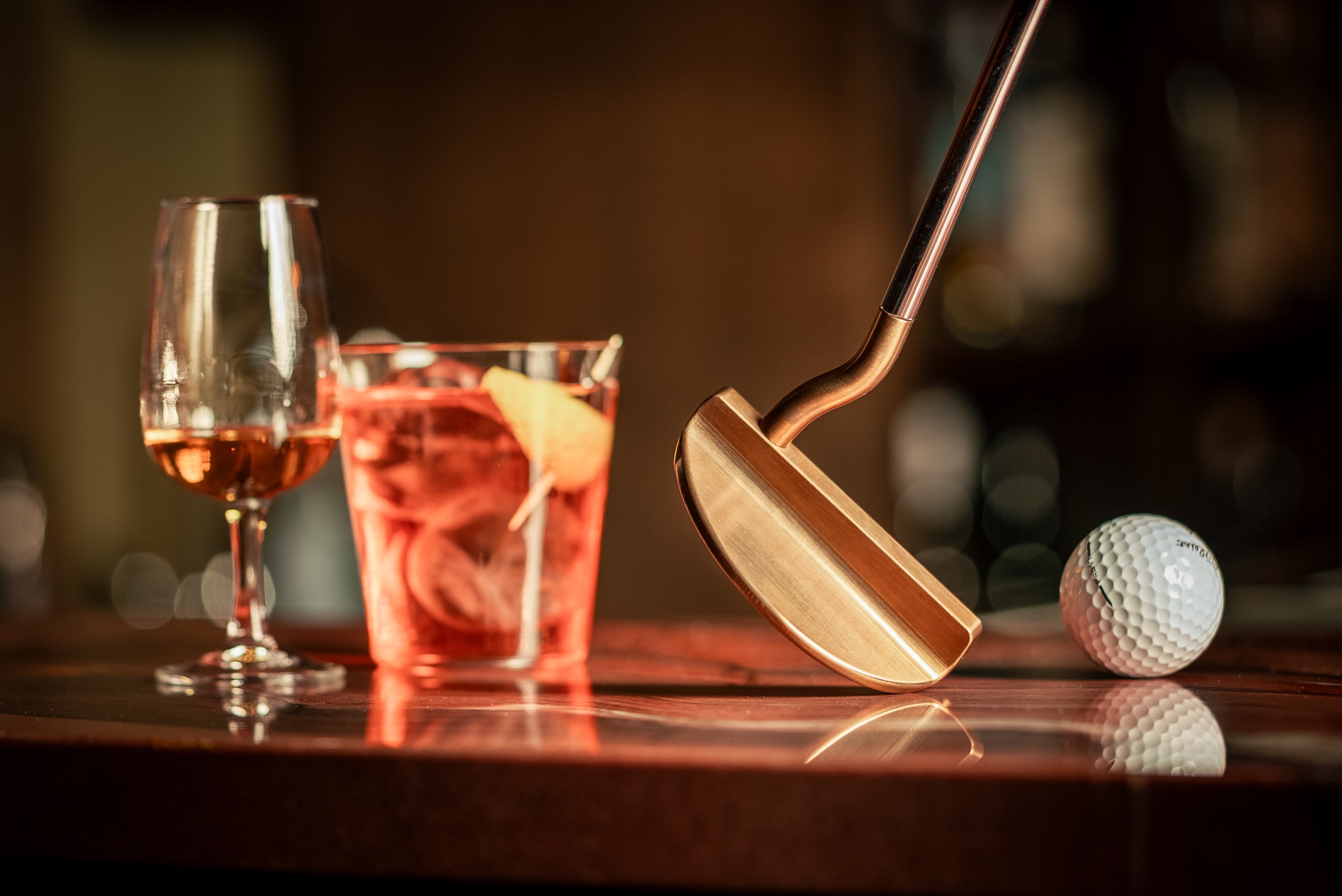 The Edel 467, a swan-neck mallet in torched gold patina, on a bar top beside two drinks and a ball