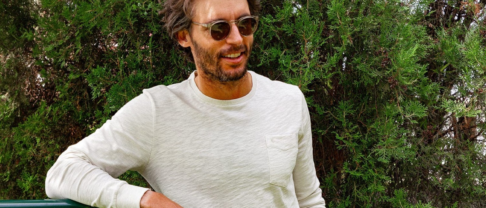 A man in Criquet’s natural slub long-sleeve pocket tee leaning on a green rail in front of cypress trees