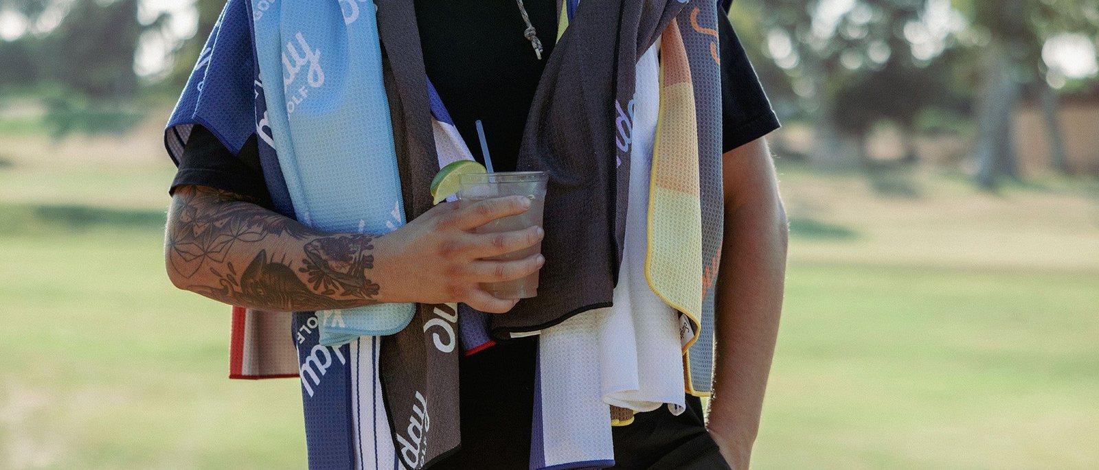 A golfer on the course with a stack of Sunday Golf towels draped over his shoulders and a drink in his hand