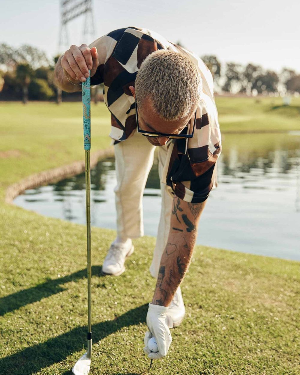 A golfer in a patterned shirt teeing up a ball beside a pond