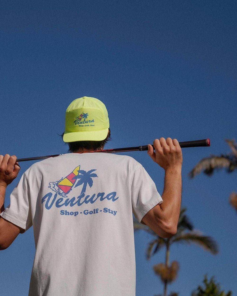 A golfer in a Ventura tee holding a club across his shoulders under palm trees