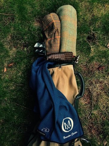Canvas and leather Sunday bag laid down in rough grass with a navy towel clipped on