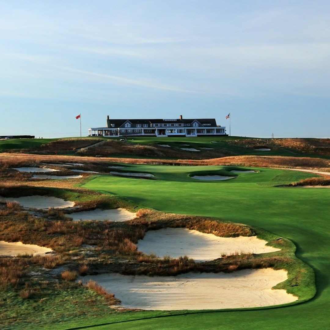 Shingled clubhouse on the hill above a green ringed by sandy waste bunkers