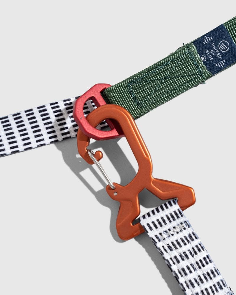 Orange carabiner clipped through a woven green webbing belt on a pale background
