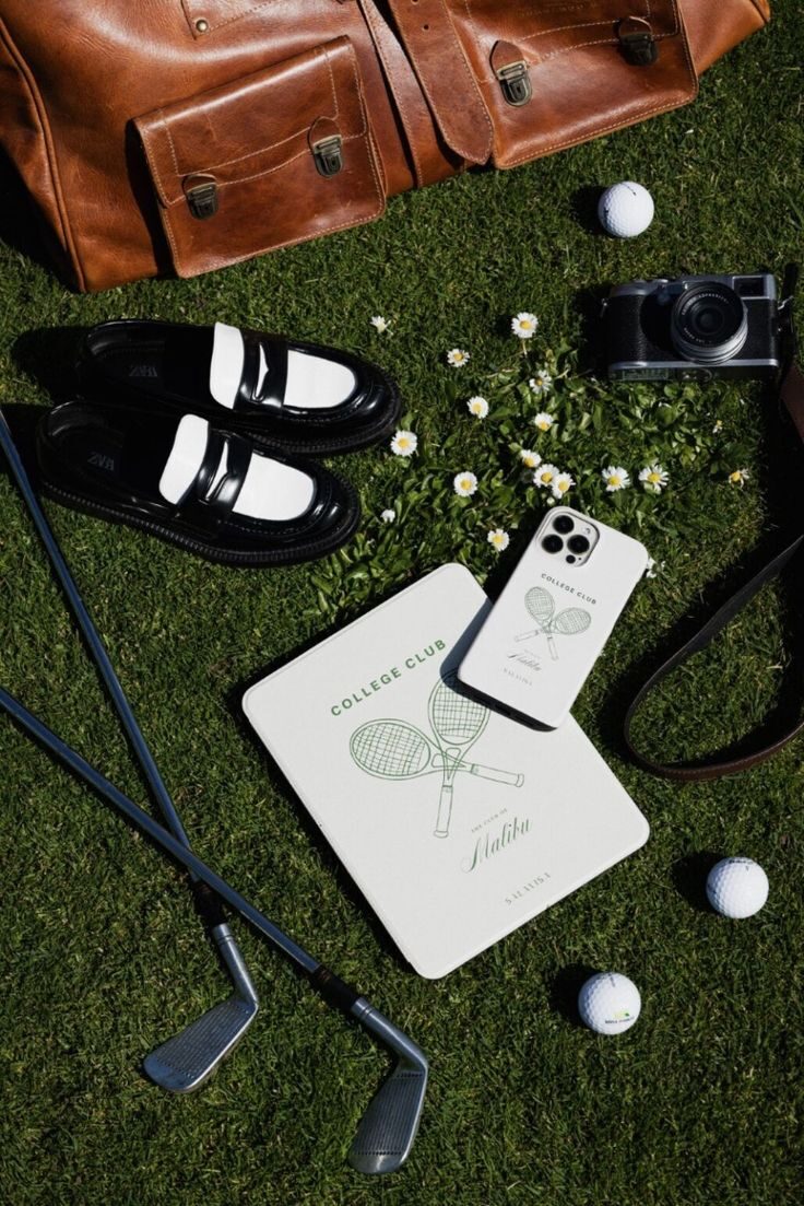 Golf lifestyle flat lay on grass with leather bag, black shoes, irons, film camera, and golf balls