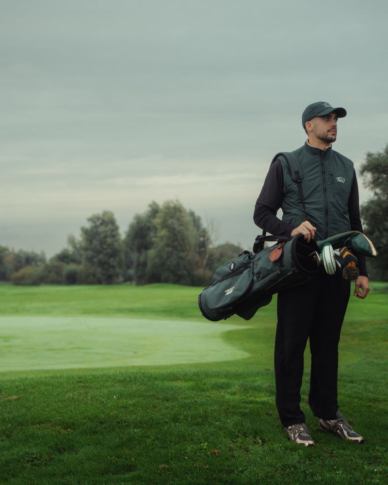 A golfer carrying a stand bag in Fella's green bodywarmer beside a flag