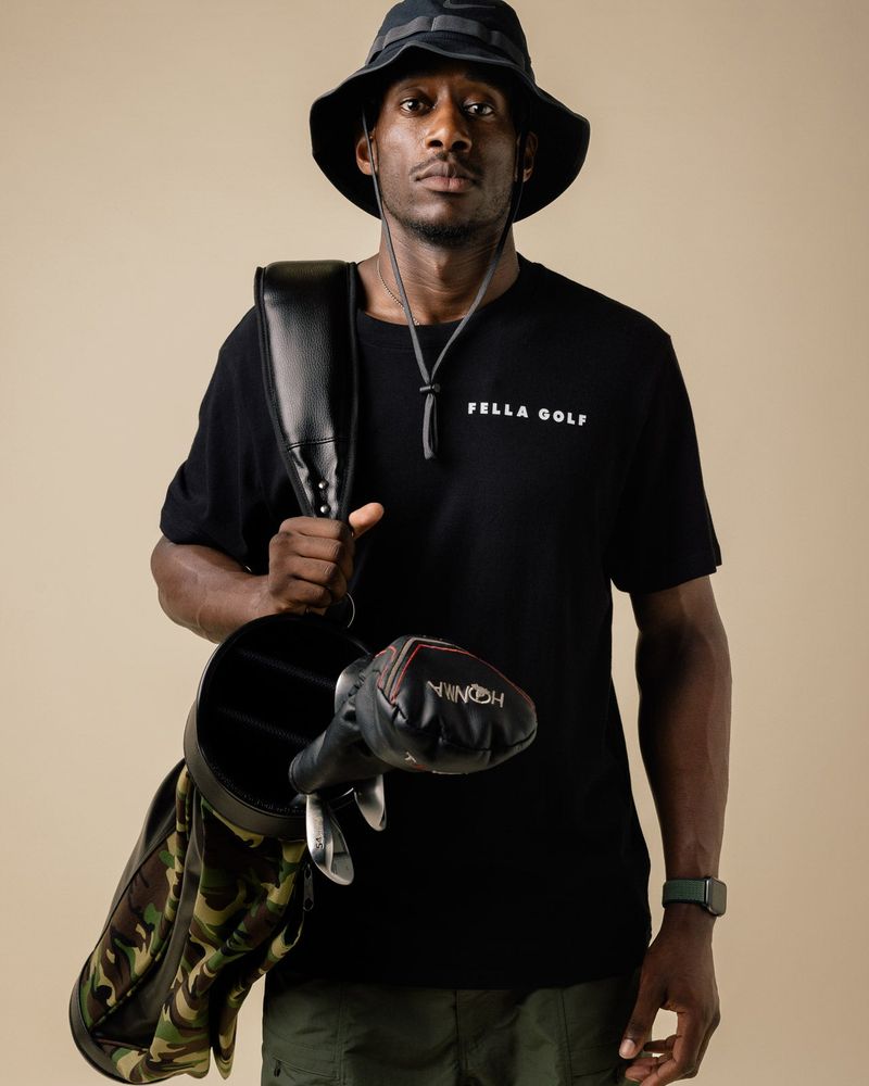 A model in the black Fella T-shirt and a bucket hat carrying a golf bag