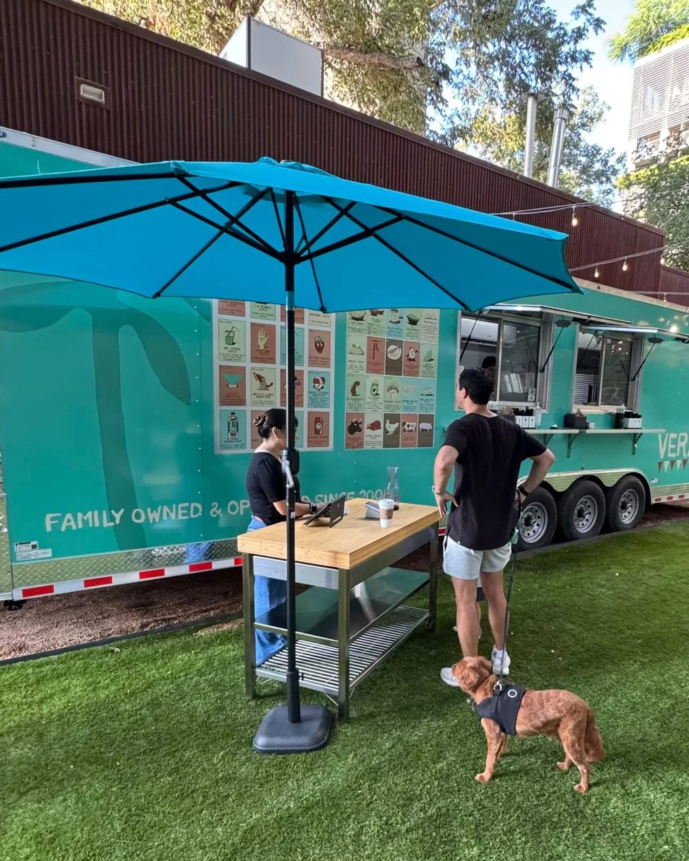 The Veracruz All Natural trailer under a blue umbrella, with a customer and a dog on the grass