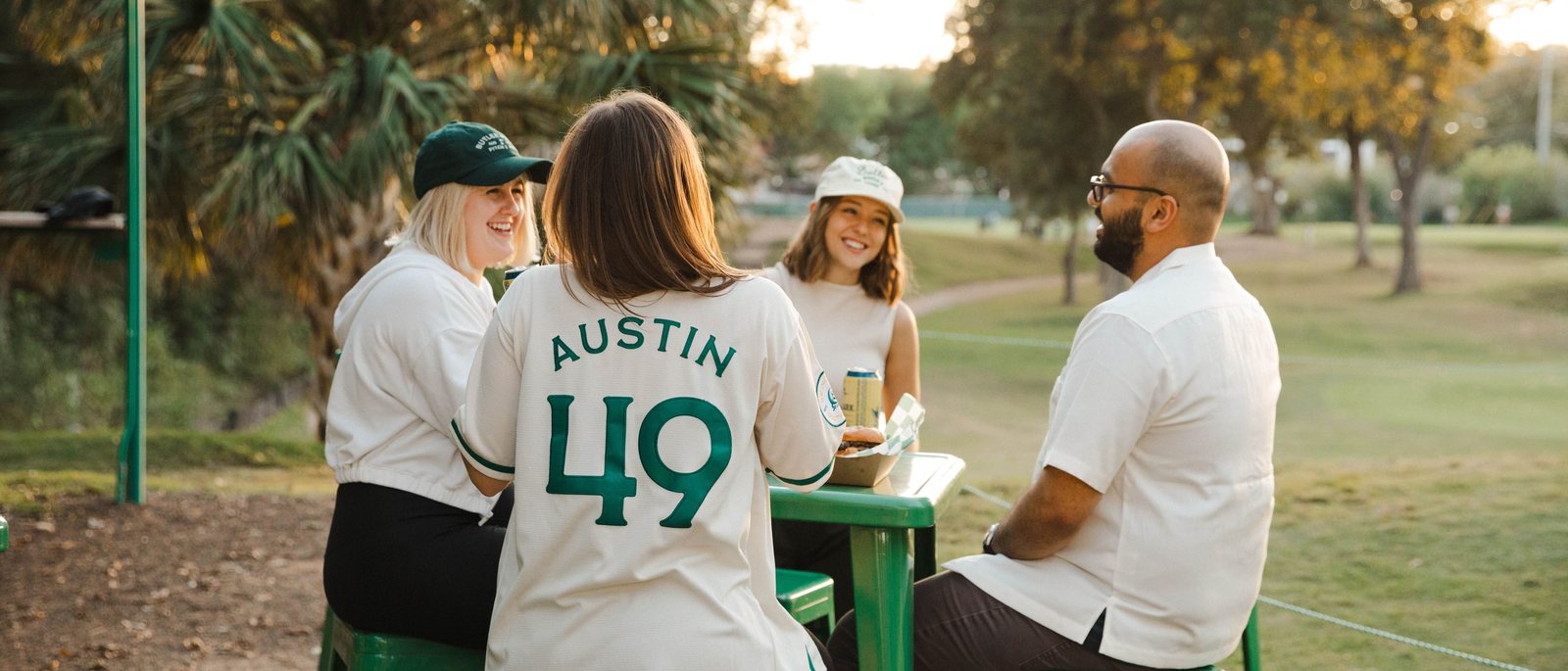 Friends at a green picnic table at Butler Pitch & Putt, one in an Austin 49 jersey, with the course behind them