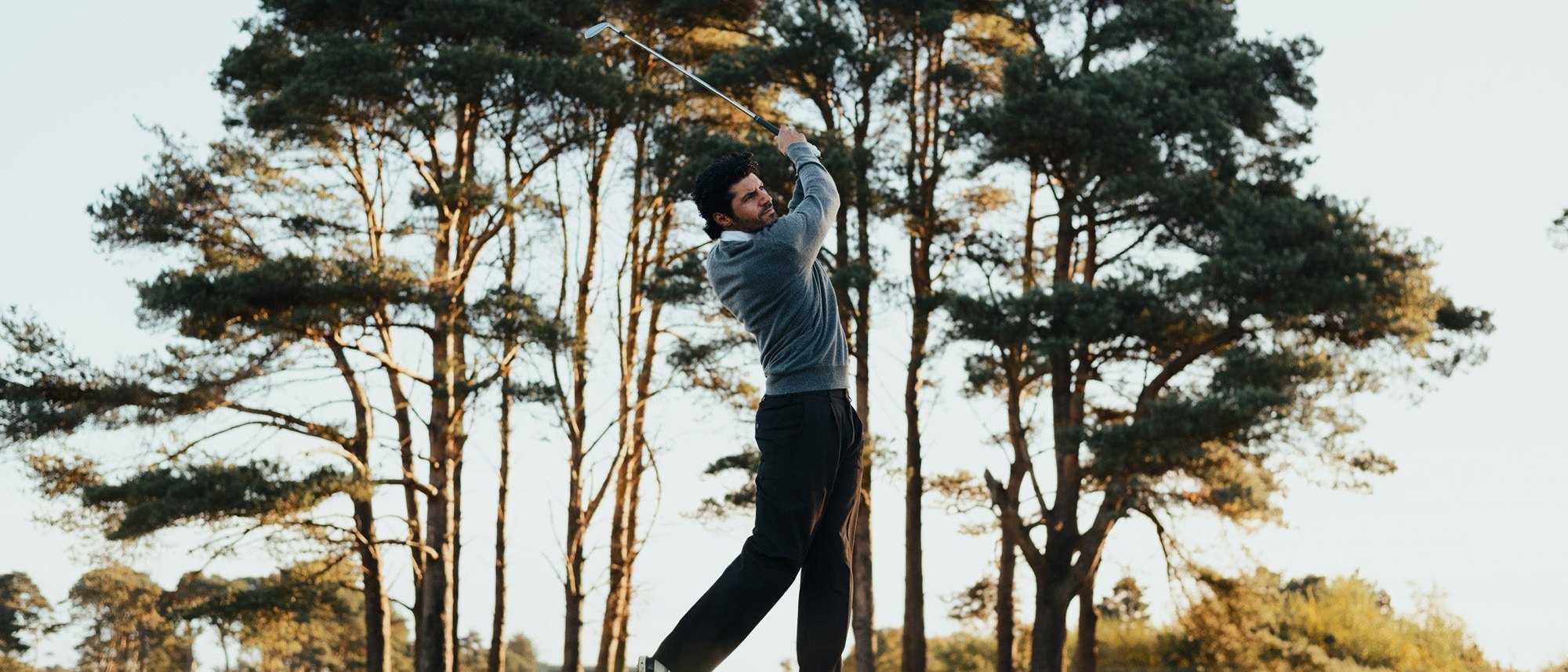 A golfer finishing his swing beneath tall Scots pines in low autumn light, from Fyfe Golf’s Golden Restless Age lookbook