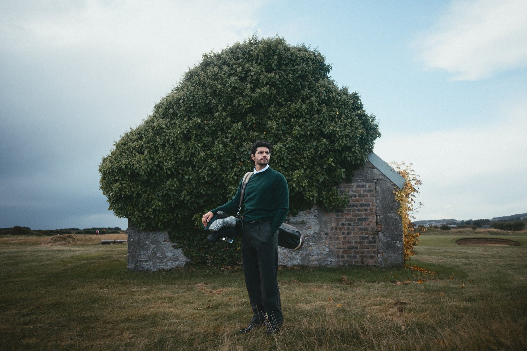 A golfer standing beside a stone hut under a huge bush on a links course