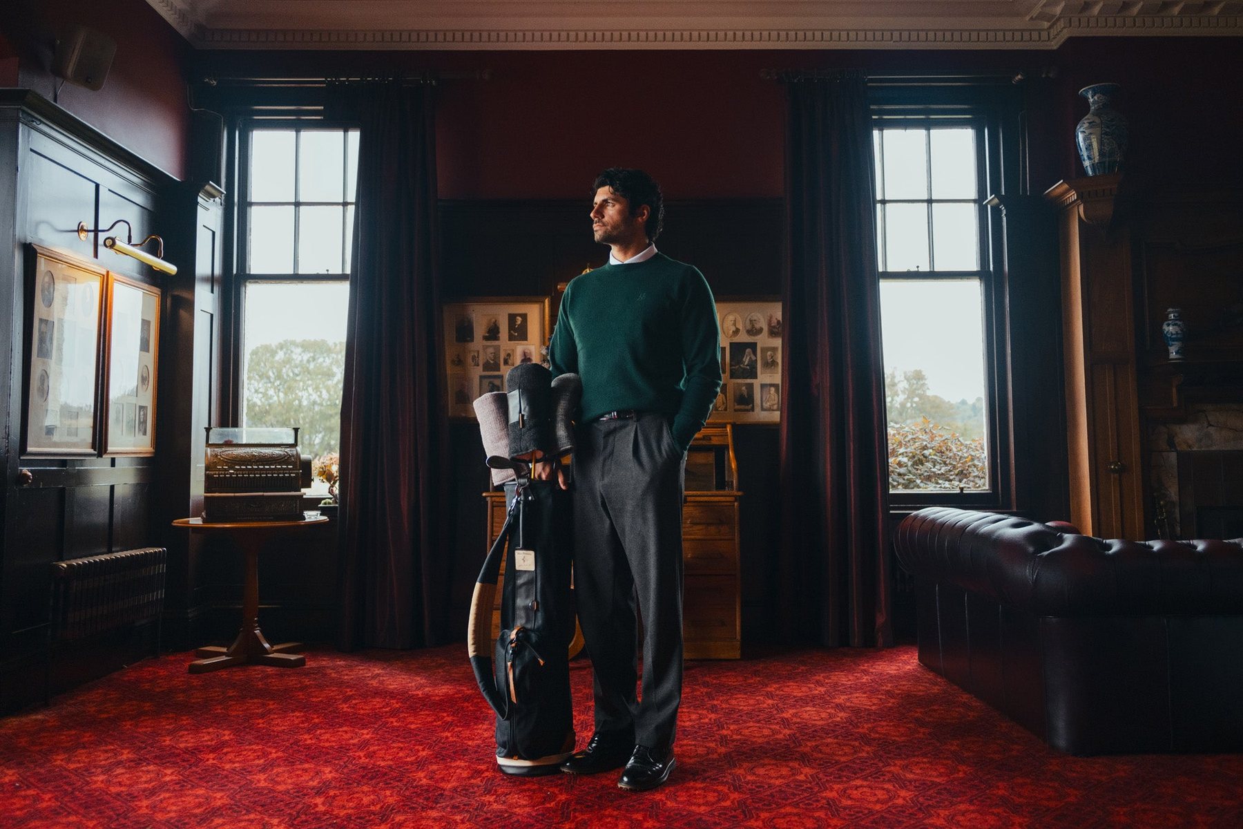 A man in a green crew neck standing by his bag in a red-carpeted clubhouse dining room