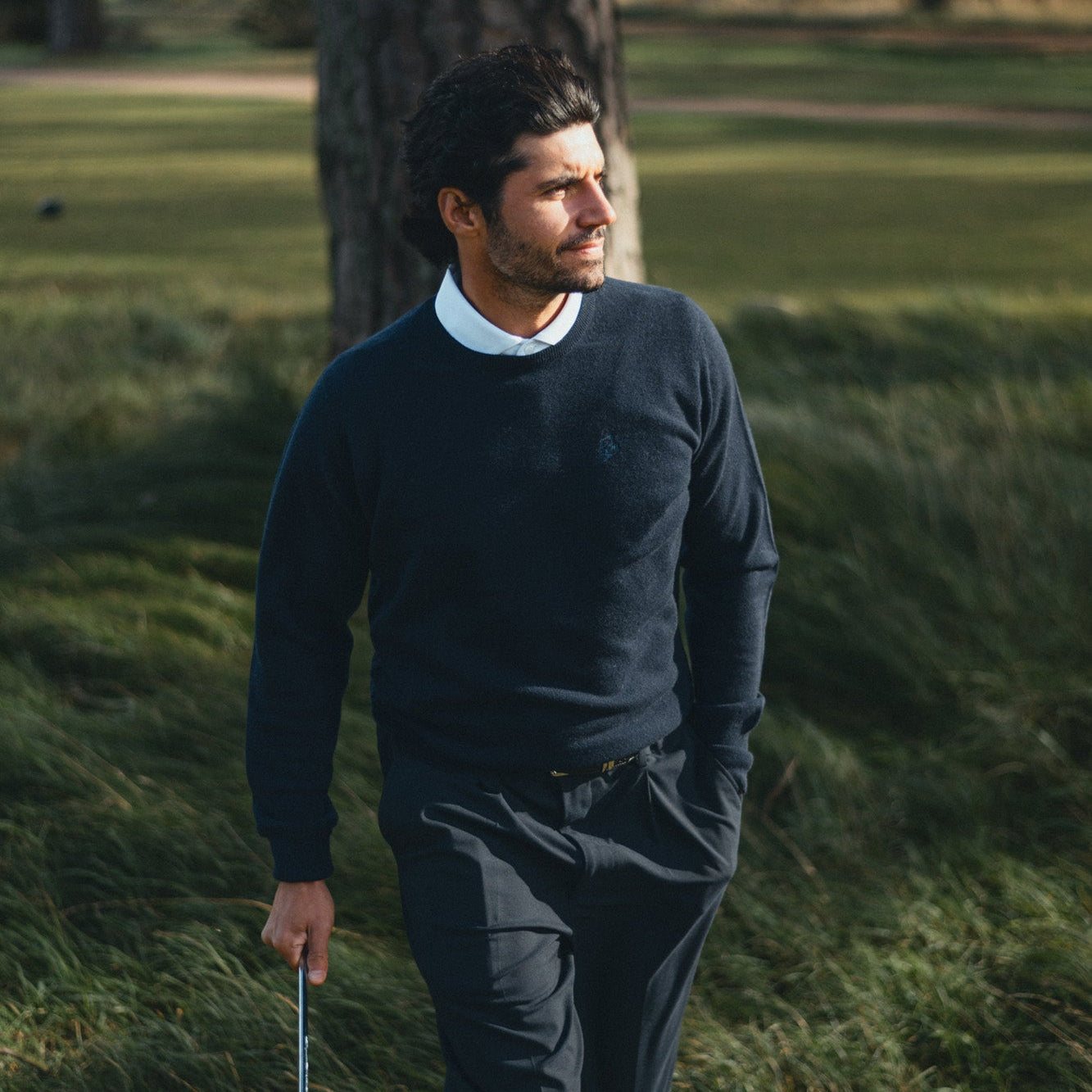 A man in a navy crew neck walking a fairway under pines