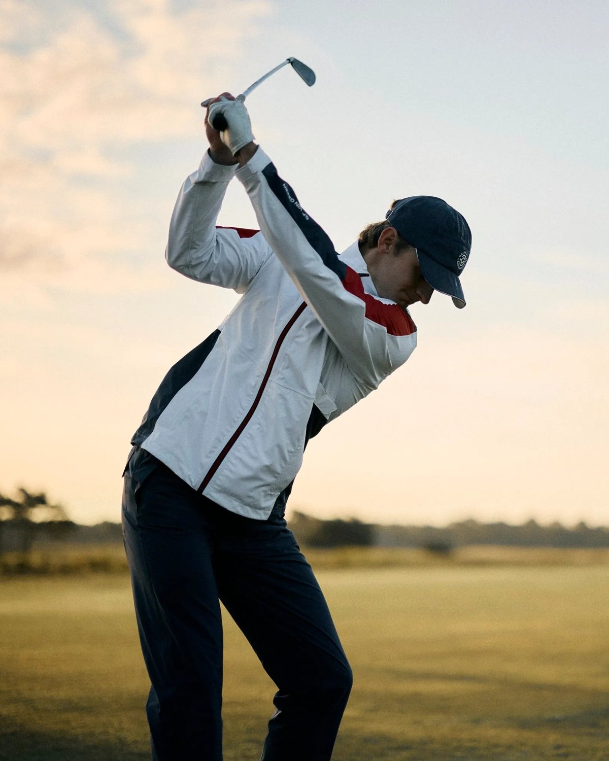 A golfer at the top of his backswing at dusk in a white and red Galvin Green jacket