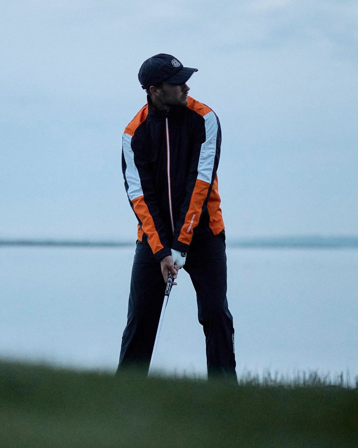 A golfer at address beside the water in a navy and orange Galvin Green jacket