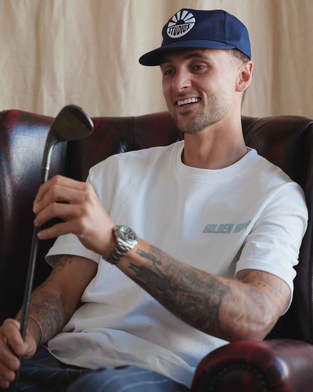 A man in a white Golden Hour tee sitting in a leather armchair holding a club