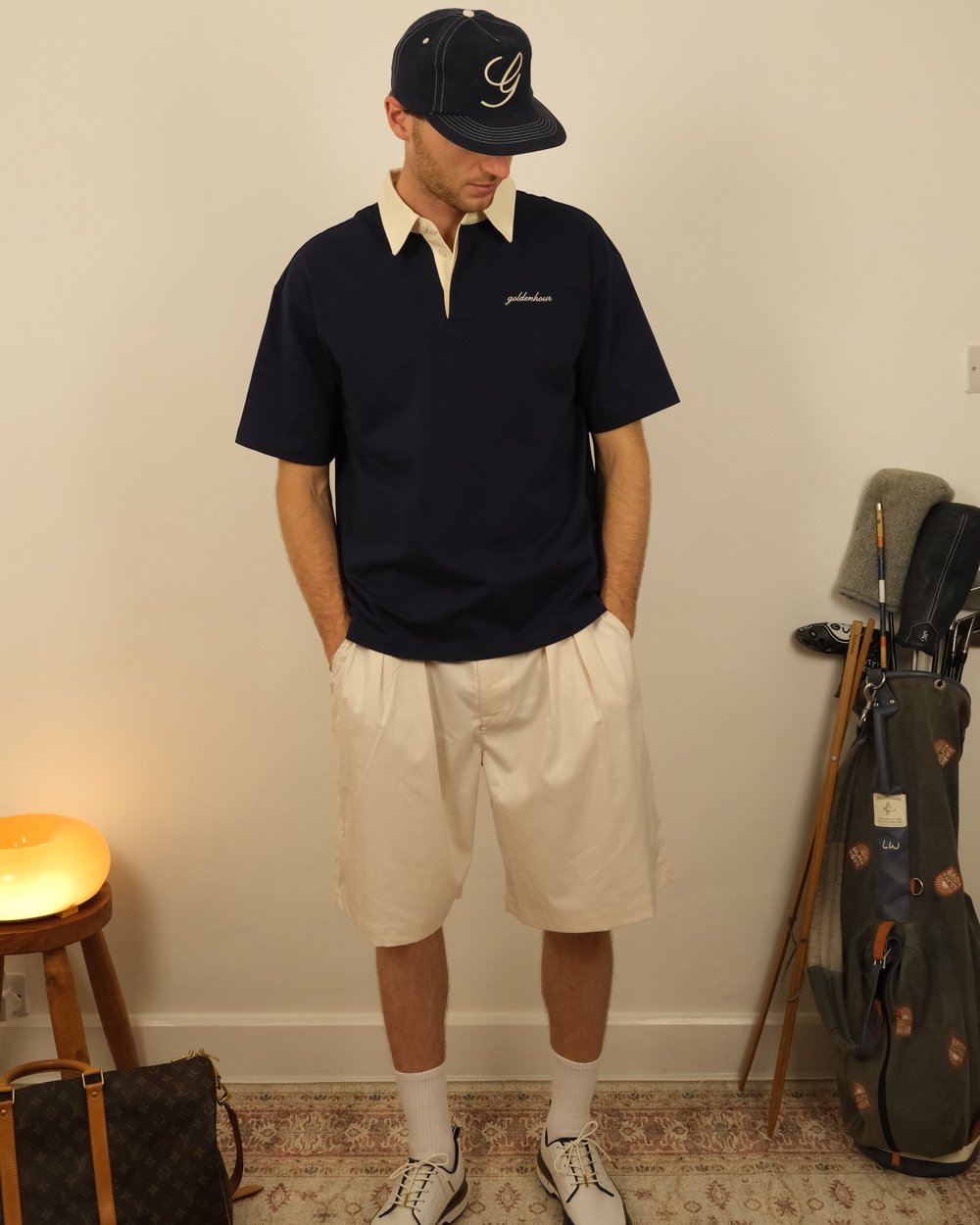 A golfer in the navy Heritage Polo and pleated shorts standing in a clubhouse beside golf bags
