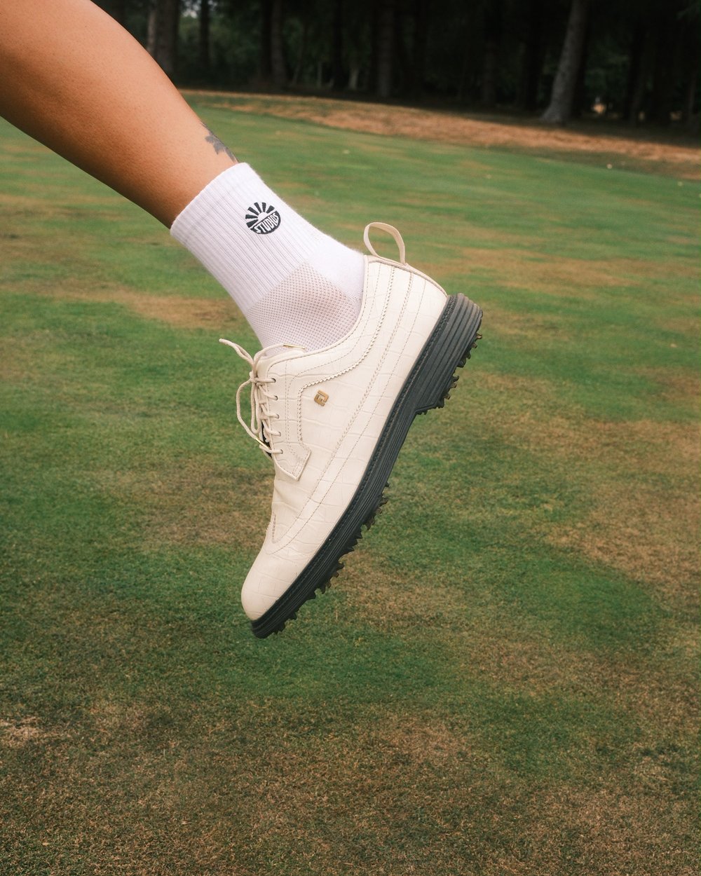 A foot in a white crew sock and golf shoe mid-stride on the grass