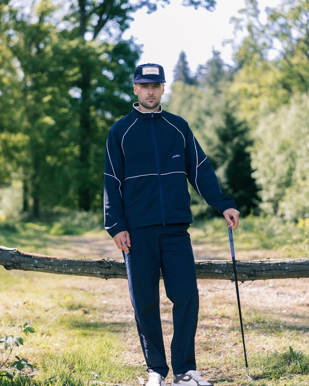 A golfer in the navy track jacket and trousers leaning on a club by a fallen tree