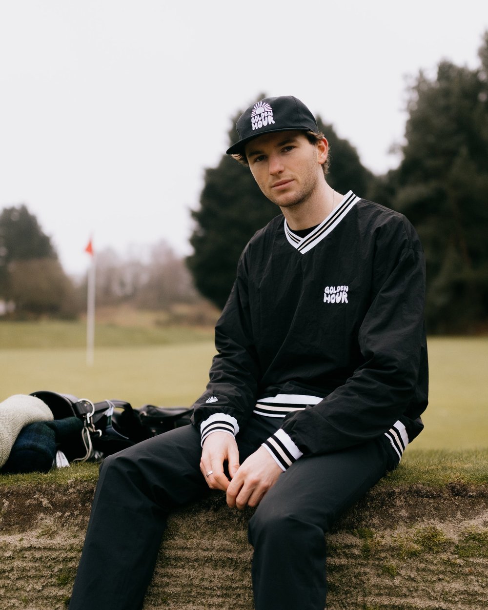 A golfer in a black windbreaker sitting by his bag near a flag